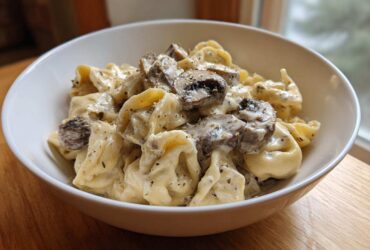A close-up of a white bowl filled with Creamy Mushroom Tortellini Alfredo, featuring tortellini coated in rich sauce and topped with sliced mushrooms.