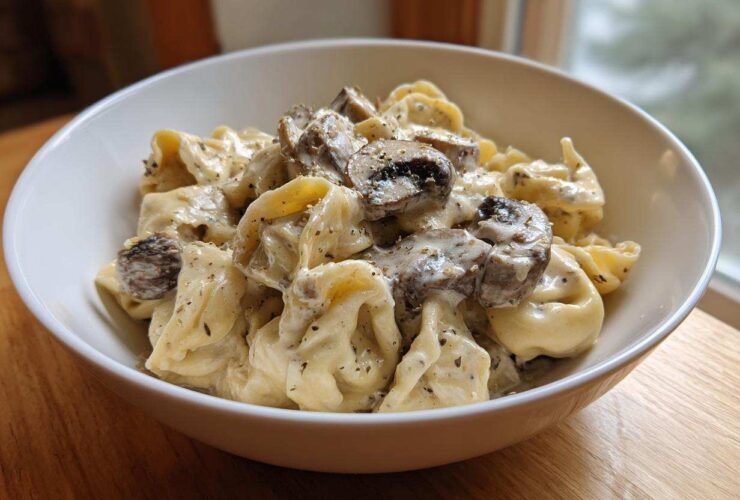 A close-up of a white bowl filled with Creamy Mushroom Tortellini Alfredo, featuring tortellini coated in rich sauce and topped with sliced mushrooms.