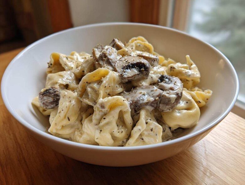 A close-up of a white bowl filled with Creamy Mushroom Tortellini Alfredo, featuring tortellini coated in rich sauce and topped with sliced mushrooms.