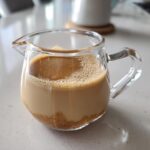 A small glass pitcher containing homemade Creme Brulee Coffee Creamer with undissolved brown sugar visible at the bottom.