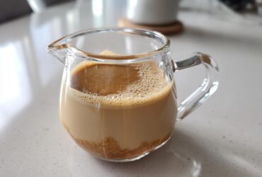 A small glass pitcher containing homemade Creme Brulee Coffee Creamer with undissolved brown sugar visible at the bottom.