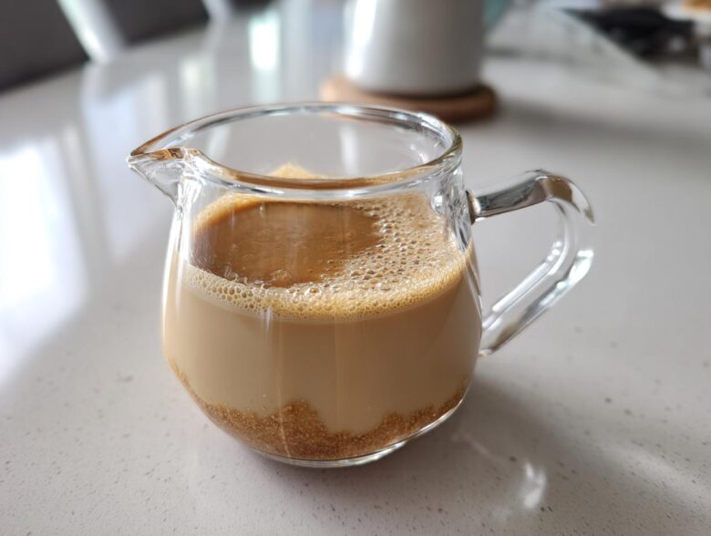 A small glass pitcher containing homemade Creme Brulee Coffee Creamer with undissolved brown sugar visible at the bottom.