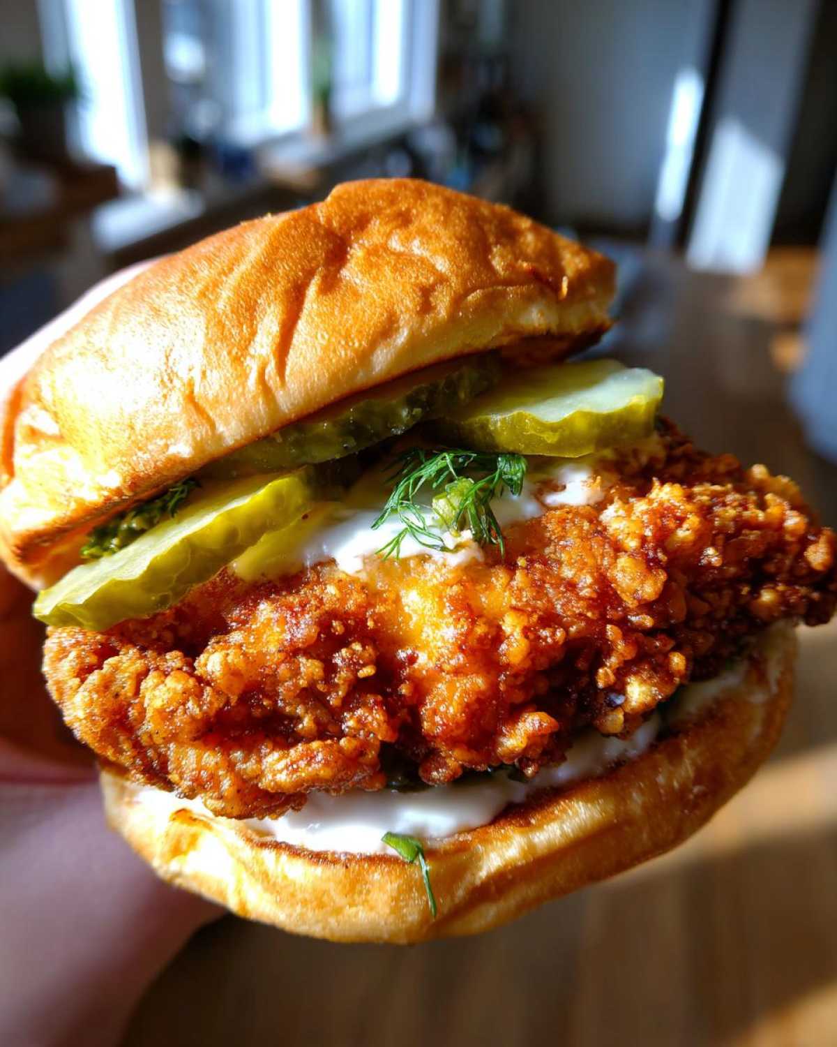 Close-up of a hand holding a delicious Crispy Chicken Sandwich featuring a thick, golden-brown fried fillet, pickles, and white sauce.