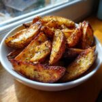 A close-up of crispy roasted Greek Lemon Potatoes seasoned with herbs in a white bowl.