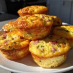 A pile of golden brown, fluffy Protein Egg Bites with visible pieces of ham and herbs on a white plate.