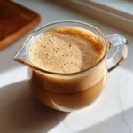 Close-up of rich, frothy Creme Brulee Coffee Creamer in a small glass pitcher on a white marble surface.