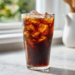 A tall, sweating glass filled with ice cubes and dark Cold Brew Coffee sitting on a marble counter.