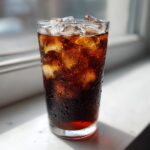 A tall, condensation-covered glass filled with ice and dark Cold Brew Coffee, sitting on a bright windowsill.