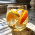 A close-up of a glass filled with ice, liquid, and slices of orange and lemon for White Peach Sangria.
