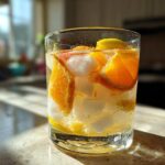A glass filled with ice, liquid, and slices of orange and lemon, representing White Peach Sangria, sitting in sunlight.