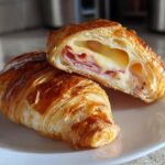 Two Golden Baked Ham and Cheese Croissants on a plate, one cut in half showing melted cheese and ham filling.