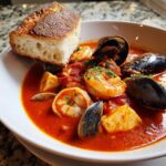 A close-up of a bowl of rich, red seafood Cioppino stew filled with shrimp, mussels, clams, and fish chunks, topped with parsley and served with crusty bread.