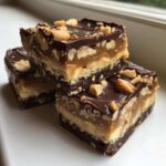 Three stacked squares of rich Snickers Fudge showing distinct layers of chocolate, nougat, caramel, and peanuts on top.