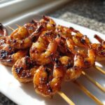 Close-up of glazed Honey Garlic Grilled Shrimp skewers piled on a white plate, showing caramelized edges.