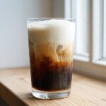 A tall glass of iced Vanilla Latte with dark coffee, ice, and a thick layer of light foam on top.
