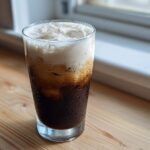 A tall glass of iced Vanilla Latte with rich, dark coffee, ice cubes, and a thick layer of white foam.