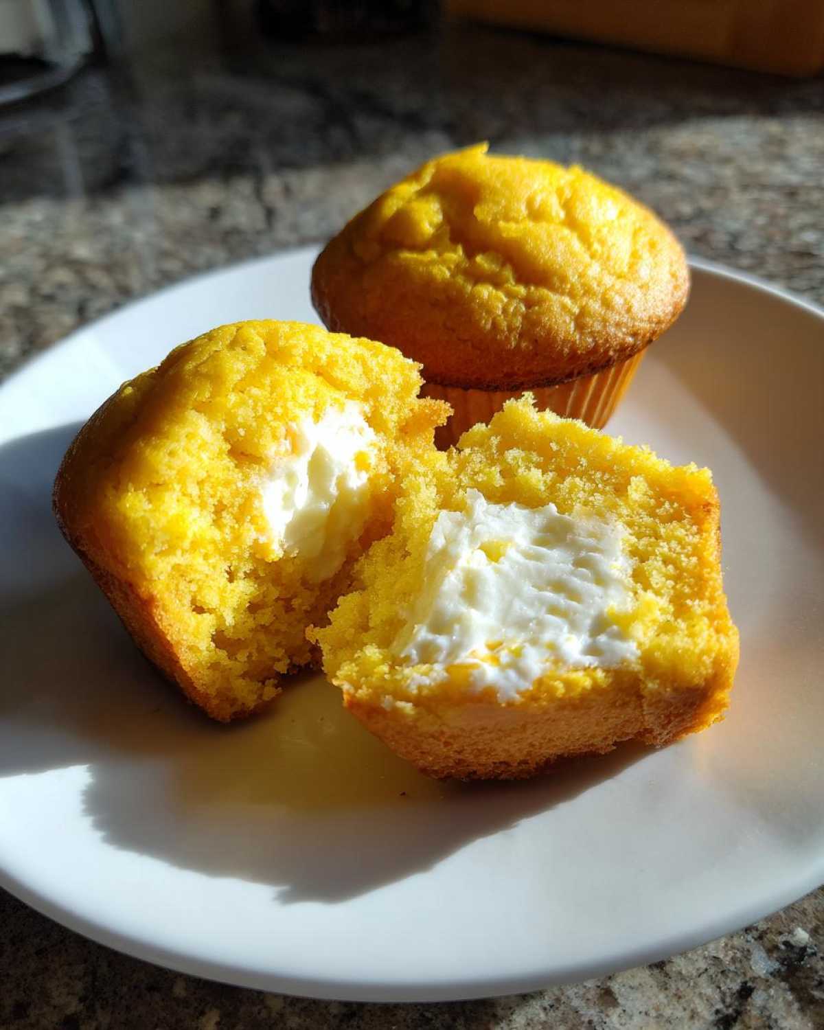 A soft lemon muffin cut in half revealing a rich cream cheese filling, next to another whole muffin.