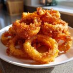 A close-up of a generous pile of golden-brown, extra crispy onion rings stacked high on a white plate.