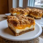 A close-up of a decadent Peach Cobbler Cheesecake Bars slice featuring a graham cracker crust, creamy cheesecake layer, peach topping, and crumble.