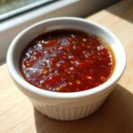 A small white ramekin filled with thick, dark red Pineapple Jalapeno Bourbon BBQ Sauce, showing visible flecks of ingredients.