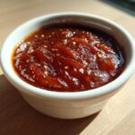 A small white ramekin filled with thick, glossy Pineapple Jalapeno Bourbon BBQ Sauce, showing visible chili flakes.