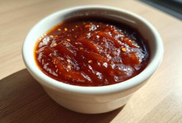A small white ramekin filled with thick, glossy Pineapple Jalapeno Bourbon BBQ Sauce, showing visible chili flakes.