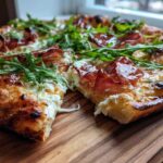 A close-up view of a slice of Prosciutto Honey Burrata Pizza showing melted burrata, prosciutto, and fresh arugula.