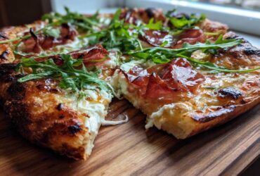 A close-up view of a slice of Prosciutto Honey Burrata Pizza showing melted burrata, prosciutto, and fresh arugula.