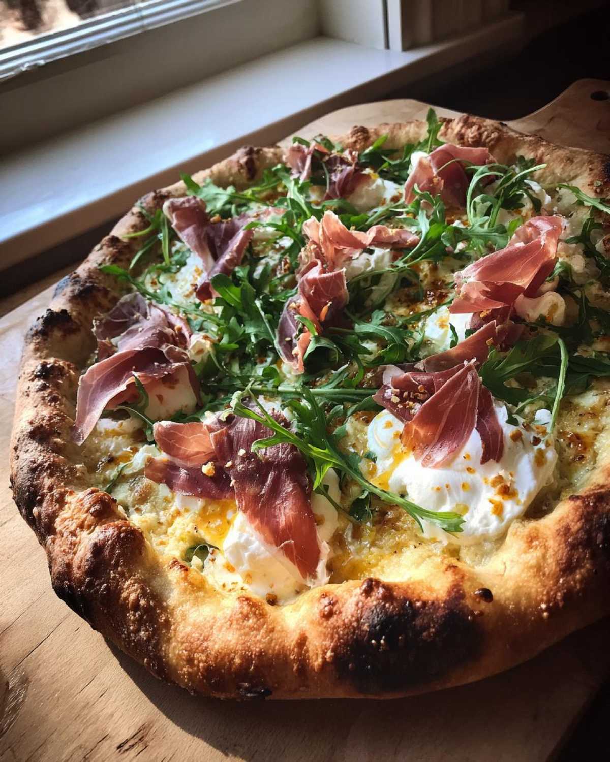 A freshly baked Prosciutto Honey Burrata Pizza topped with prosciutto, creamy burrata, and fresh arugula, resting on a wooden peel.