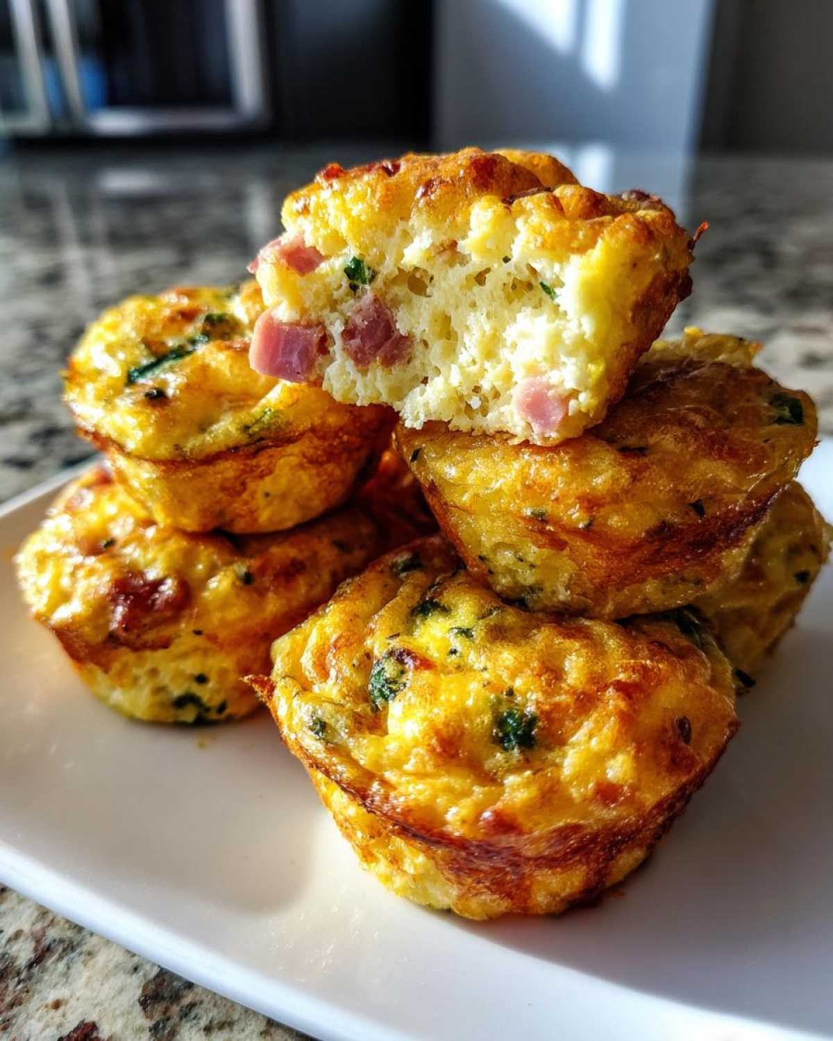 A stack of fluffy Protein Egg Bites with visible ham and green herbs, one bite is broken open to show the texture.