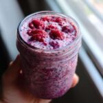 A hand holding a glass jar filled with vibrant pink Raspberry Chia Seed Pudding, topped with fresh raspberries.