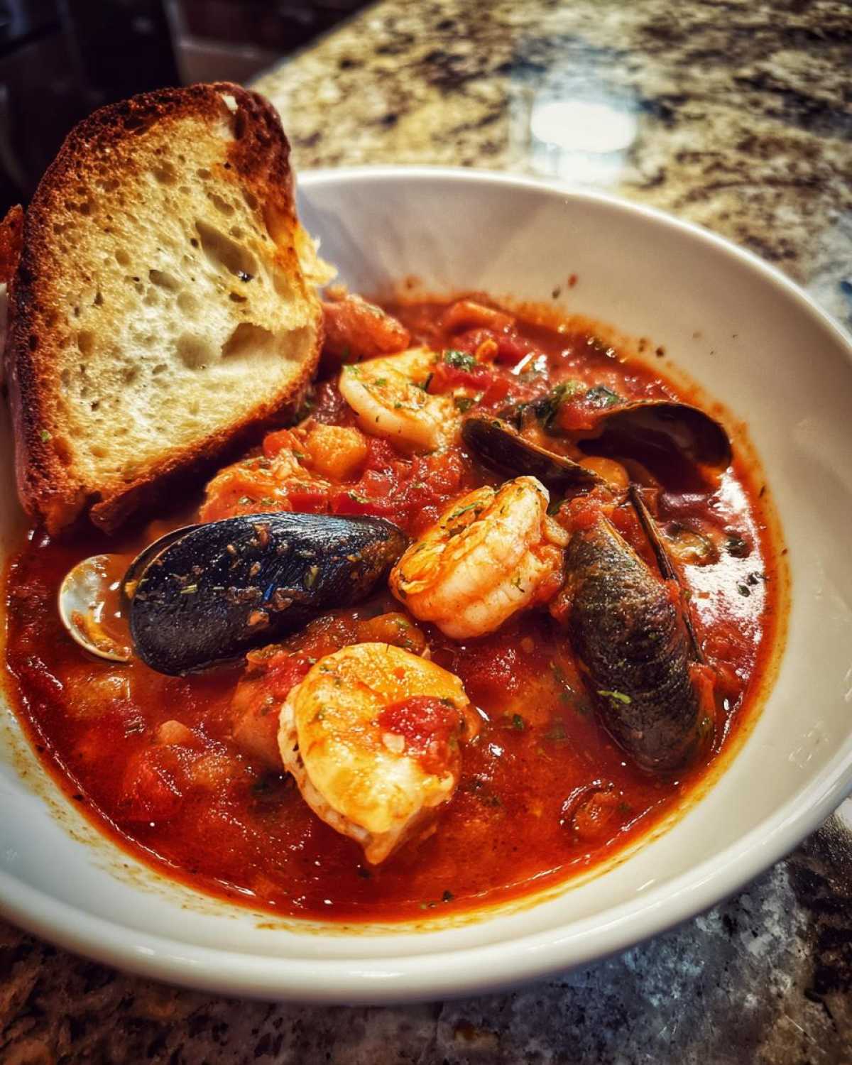 A close-up of a white bowl filled with rich, red seafood Cioppino stew featuring shrimp and mussels, served with a slice of toasted bread.