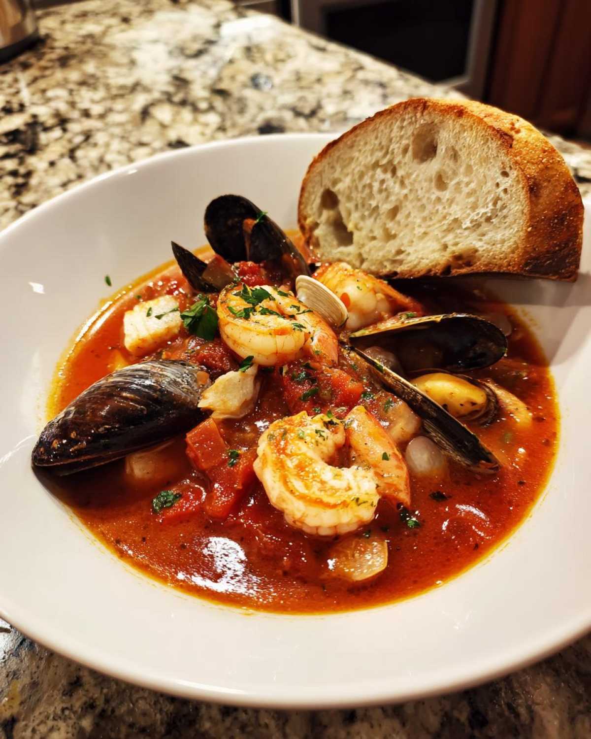 A close-up of a bowl of rich, red tomato-based Cioppino stew filled with shrimp, mussels, clams, and served with a large slice of crusty bread.