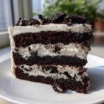 A tall slice of Cookies and Cream Cake with dark chocolate layers and white frosting, topped with cookie pieces.