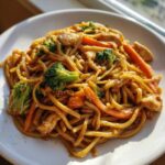 Close-up of a plate of Stir Fried Lo Mein featuring noodles, chicken pieces, bright green broccoli, and sliced carrots.