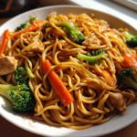 A close-up of a white bowl filled with glossy Stir Fried Lo Mein noodles, chicken pieces, bright green broccoli florets, and sliced carrots.