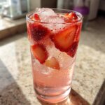 Close-up of a tall, sweating glass filled with pink Strawberry Lemonade, ice, and fresh strawberry slices.