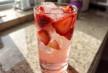 Close-up of a tall, sweating glass filled with pink Strawberry Lemonade, ice, and fresh strawberry slices.