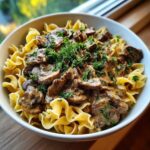 Close-up of a white bowl filled with creamy Beef Stroganoff served over wide egg noodles and topped with fresh parsley.