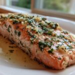 Close-up of a perfectly cooked piece of Baked Salmon topped with melted butter, minced garlic, and fresh green herbs.