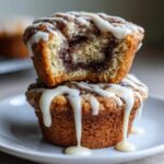 Two stacked Cinnamon Roll Muffins, one with a bite taken out showing the swirl, drizzled heavily with white icing.
