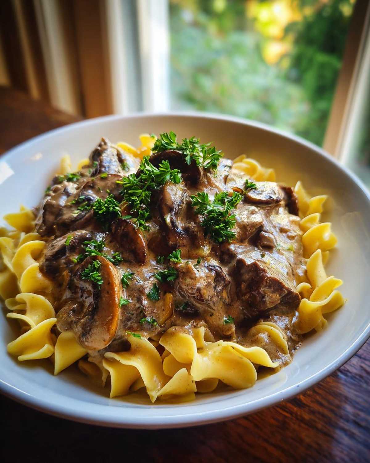 A close-up of creamy Beef Stroganoff with mushrooms and beef chunks served over wide egg noodles, garnished with parsley.