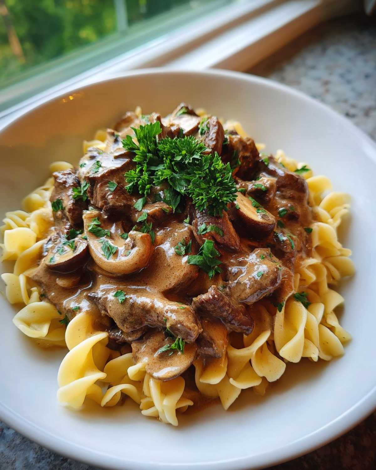 A close-up of creamy Beef Stroganoff served over wide egg noodles, garnished with fresh parsley.