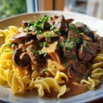 Close-up of a bowl of creamy Beef Stroganoff with tender beef chunks and mushrooms served over curly egg noodles, garnished with parsley.