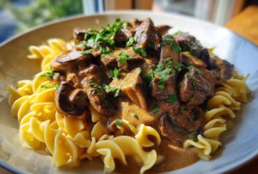 Close-up of a bowl of creamy Beef Stroganoff with tender beef chunks and mushrooms served over curly egg noodles, garnished with parsley.