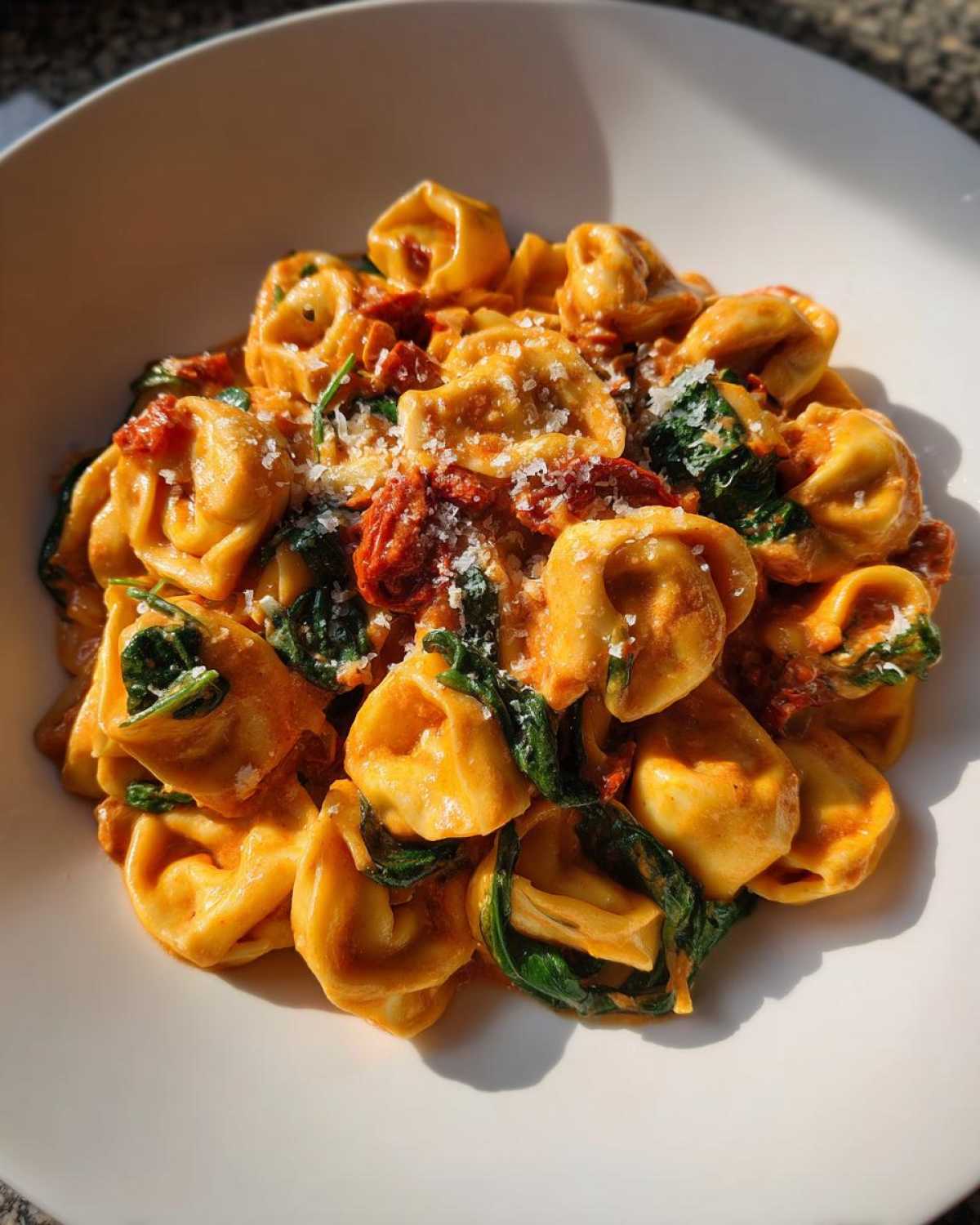 A close-up, sunlit bowl of creamy Marry Me Tortellini mixed with wilted spinach and sun-dried tomatoes, topped with grated cheese.