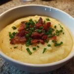 A bowl of thick, creamy potato soup topped generously with crispy bacon bits and fresh chopped chives.