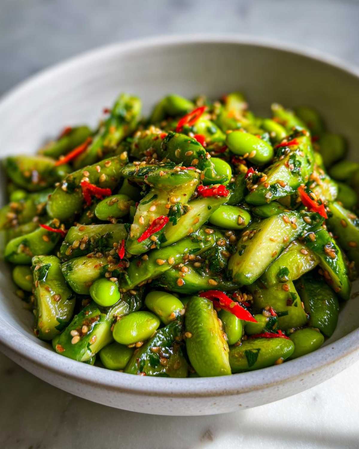 Close-up of a vibrant Edamame and Cucumber Salad tossed in a savory dressing with sesame seeds and red chili slices.