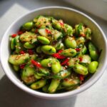Close-up of a vibrant Edamame and Cucumber Salad mixed with red chili slices and sesame seeds in a white bowl.