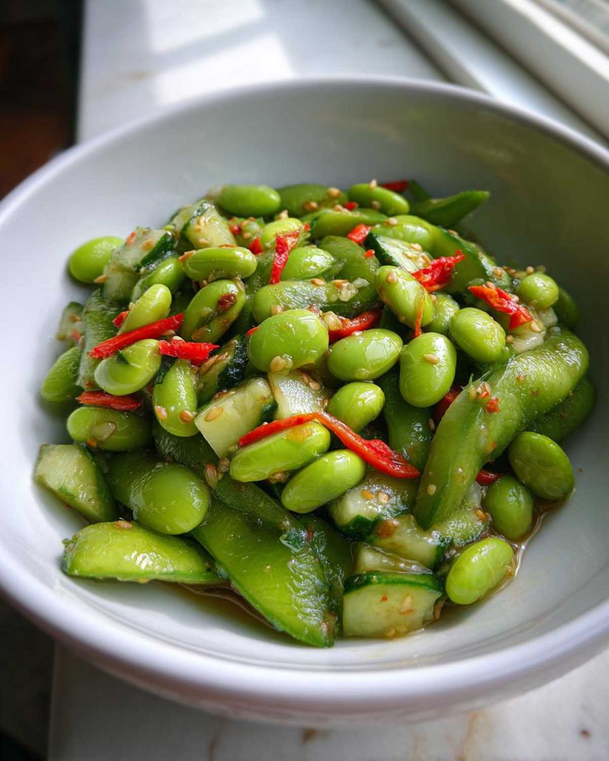 Close-up of a vibrant Edamame and Cucumber Salad mixed with red chili slices in a white bowl.